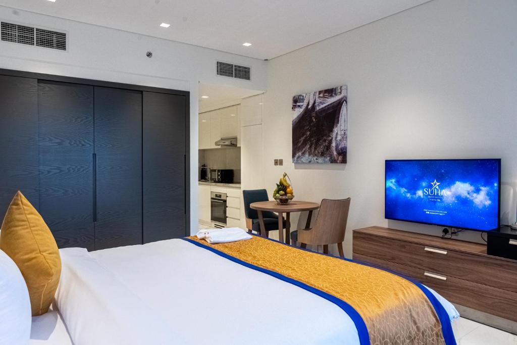 The spacious room features a large bed with a golden and blue accent cover, a flat-screen TV on a wooden cabinet, and a cozy dining area with a table and two chairs. A fully equipped kitchen is visible in the background, complemented by dark cabinetry.