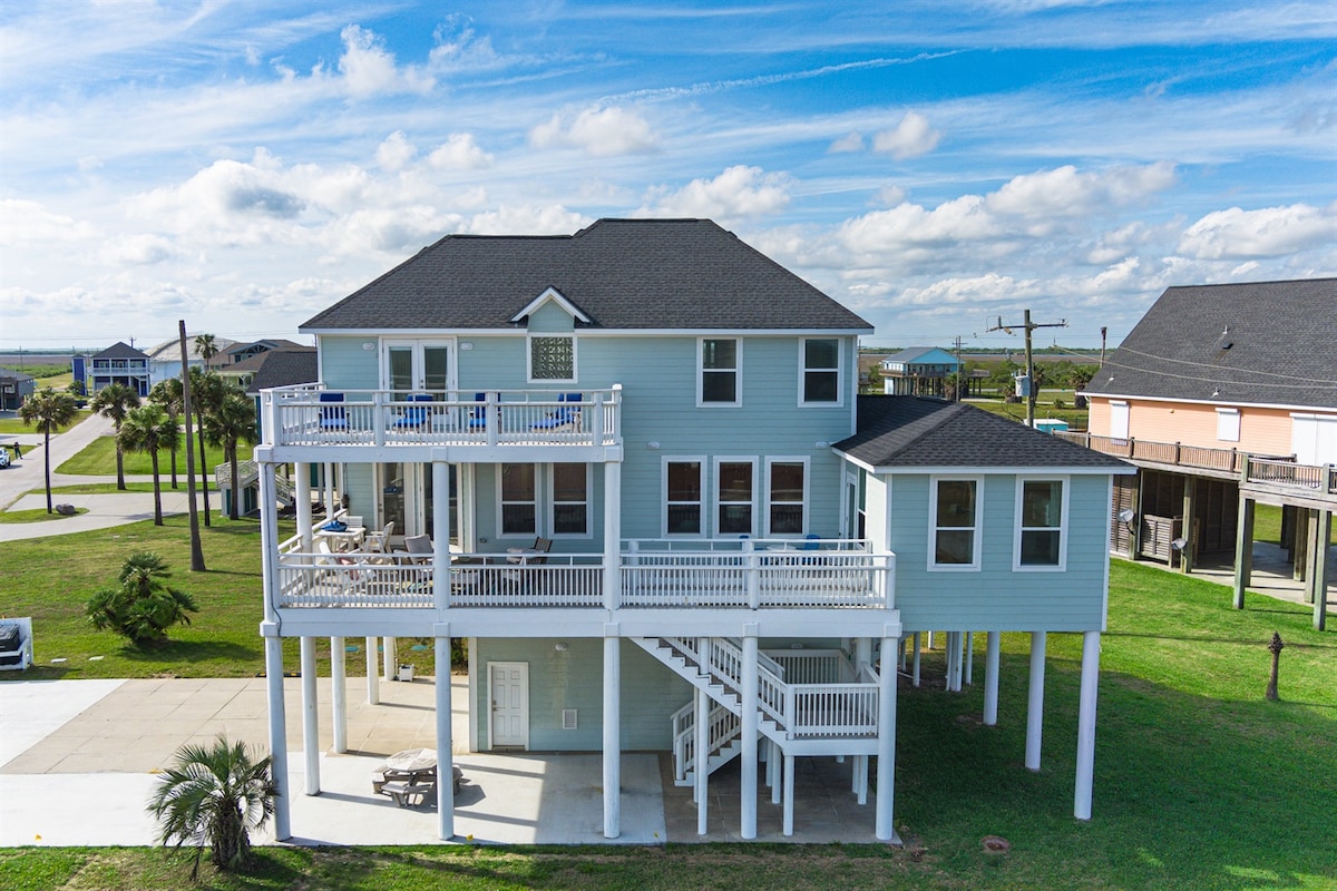 This spacious beach house features multiple decks and a vibrant exterior, surrounded by lush green grass. The property stands elevated on stilts, offering a commanding view of the neighborhood. Large windows invite natural light inside, while the nearby ocean can be glimpsed in the distance.