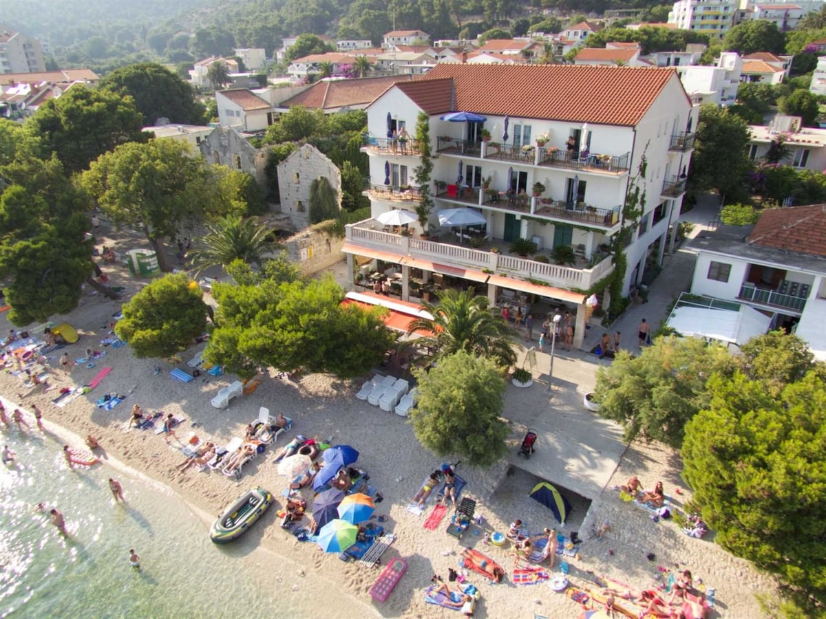 An aerial view captures a charming building located directly on a sandy beach, featuring a red-tiled roof and multiple balconies. The surrounding area is filled with beachgoers sunbathing and relaxing near the water amidst lush greenery, enhancing the serene coastal environment.