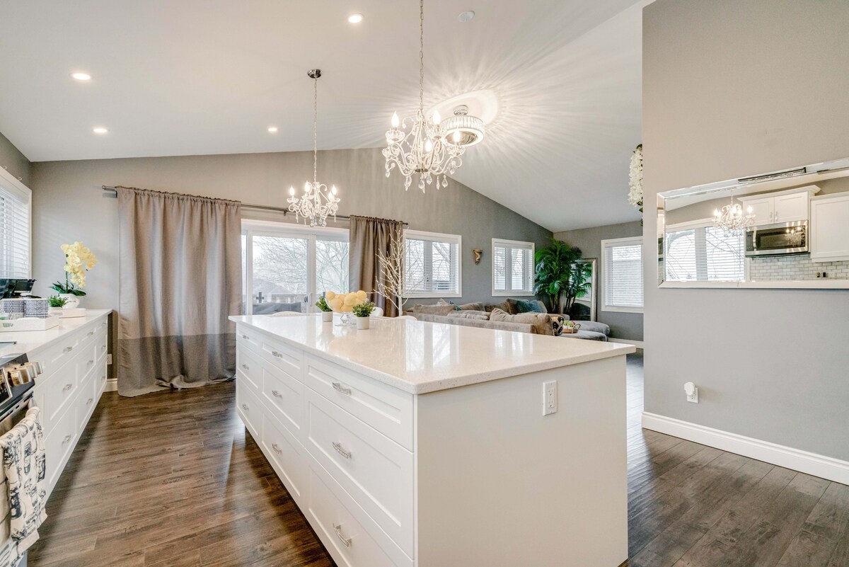 A spacious kitchen area features a large central island with a polished countertop, surrounded by ample cabinetry. Natural light filters through multiple windows dressed in soft curtains, highlighting the elegant lighting fixtures and open layout that lends a modern ambiance to the space.