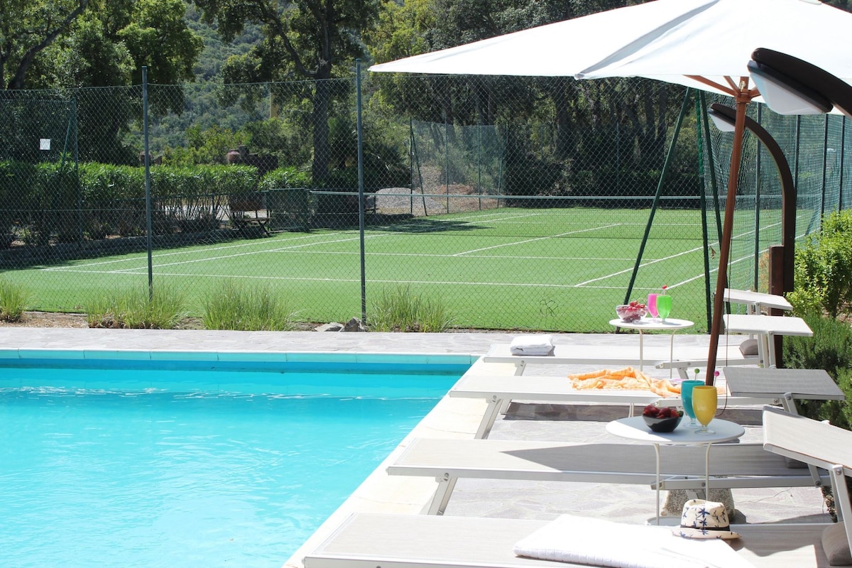 A bright swimming pool is set in the foreground, featuring lounge chairs with umbrellas nearby. In the background, a well-maintained tennis court is visible, surrounded by greenery. A small table displays refreshments, enhancing the outdoor space.