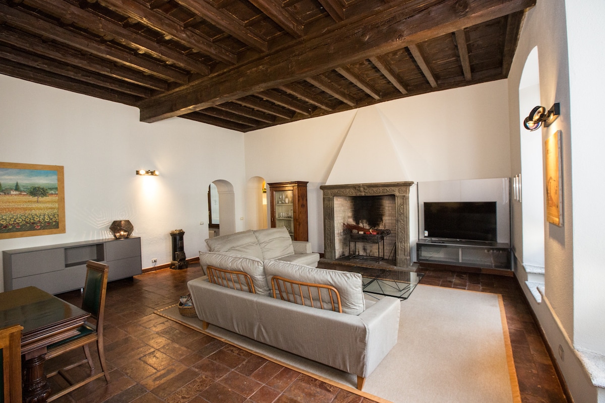 A spacious living room features high wooden beams on the ceiling and large arched windows allowing natural light. Two sofas are arranged around a glass coffee table, while a TV is positioned near a stone fireplace. Artwork and decorative pieces enhance the room's ambiance.