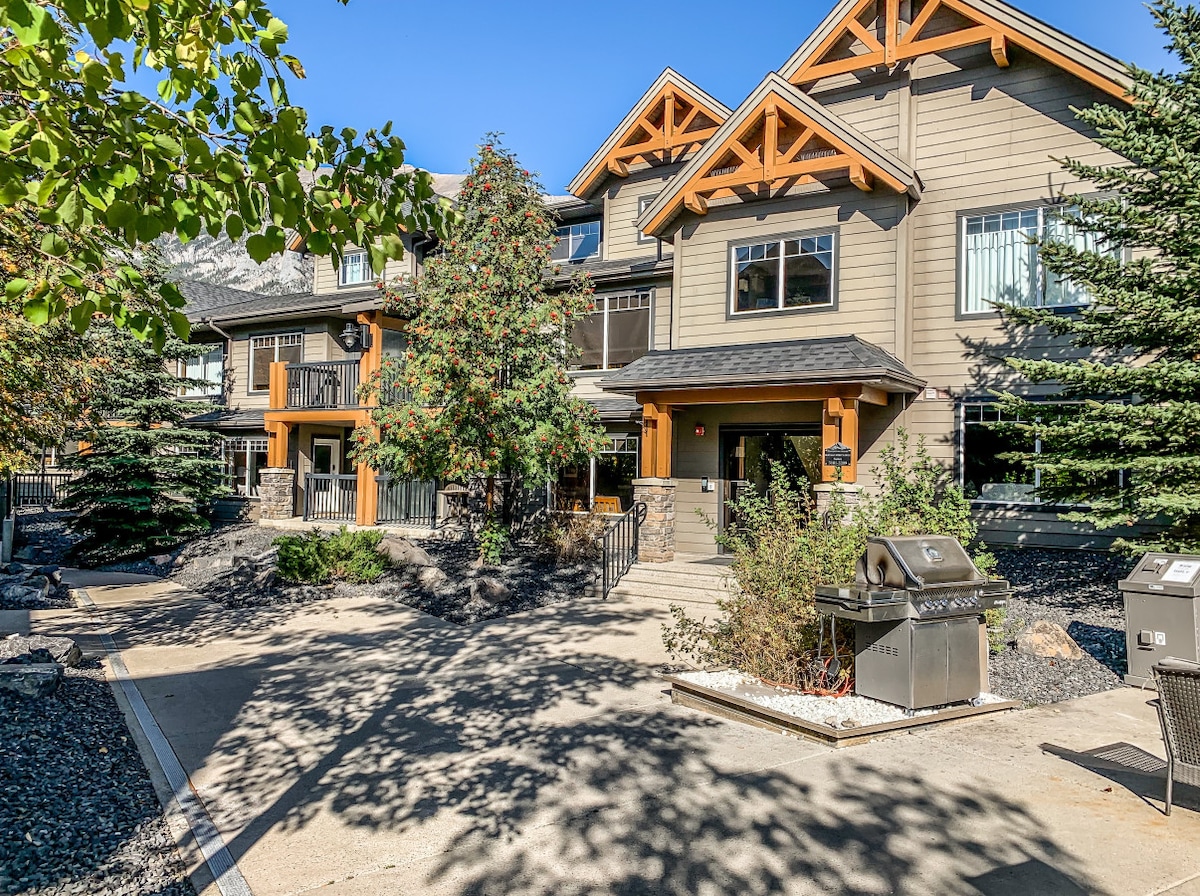 The exterior of the Copperstone Resort is shown, featuring a rustic design with wooden accents and a welcoming entryway. The surrounding area includes landscaped greenery, concrete pathways, and two barbecue grills, inviting guests to enjoy the outdoor space in a serene mountain setting.