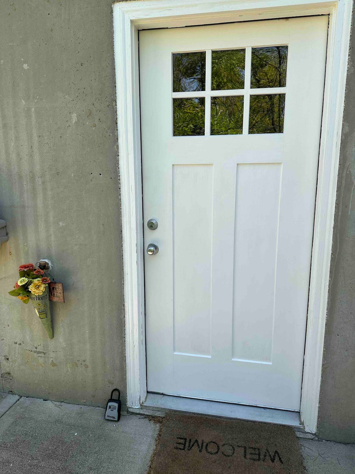 A white door with a window at the top is set against a concrete wall. A welcome mat is positioned at the entrance, and a bouquet of flowers is attached to the wall nearby. A keyless entry pad is visible on the ground beside the door.