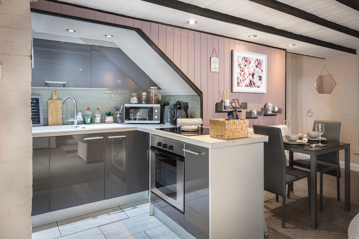 The kitchen area features modern, sleek cabinetry with a ceramic hob and multi-function oven. A dining table is set for two, complemented by elegant tableware. Ambient lighting highlights the space, including decorative wall art and a thoughtfully arranged collection of kitchen essentials.