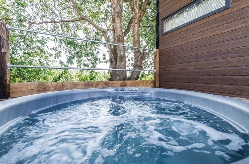 A private hot tub featuring bubbling water is positioned outdoors, bordered by wooden decking and trees. Natural light filters through the surrounding foliage, creating a serene atmosphere. The hot tub is framed by a stylish wooden structure, offering an inviting space for relaxation.