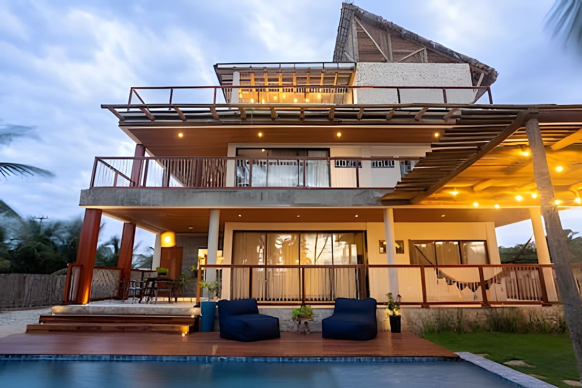 The exterior of the house reveals a multi-level structure with a spacious balcony and lounge seating. Soft lighting highlights the architectural features, while the inviting swimming pool lies in the foreground. Tropical greenery surrounds the property, enhancing the tranquil setting near the beach.