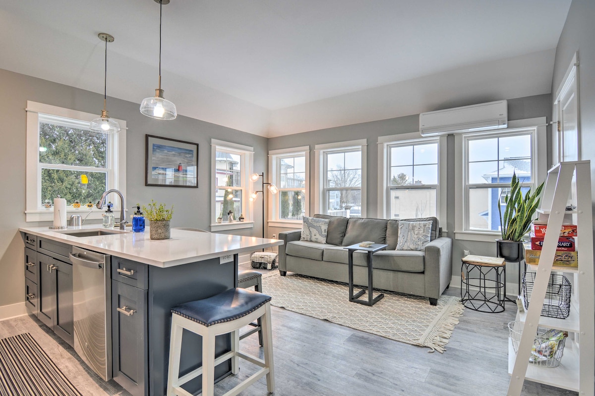 A bright living space features large windows allowing natural light to fill the room. A comfortable gray sofa is complemented by a light area rug and a coffee table. The kitchen area includes a modern island with bar seating and stainless steel appliances.