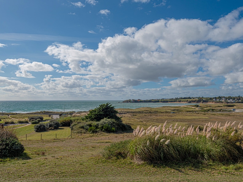 A scenic view captures the coastline of Guidel, with rolling green fields leading toward the ocean. Gentle waves can be seen under a bright sky dotted with fluffy clouds, creating a tranquil natural landscape.