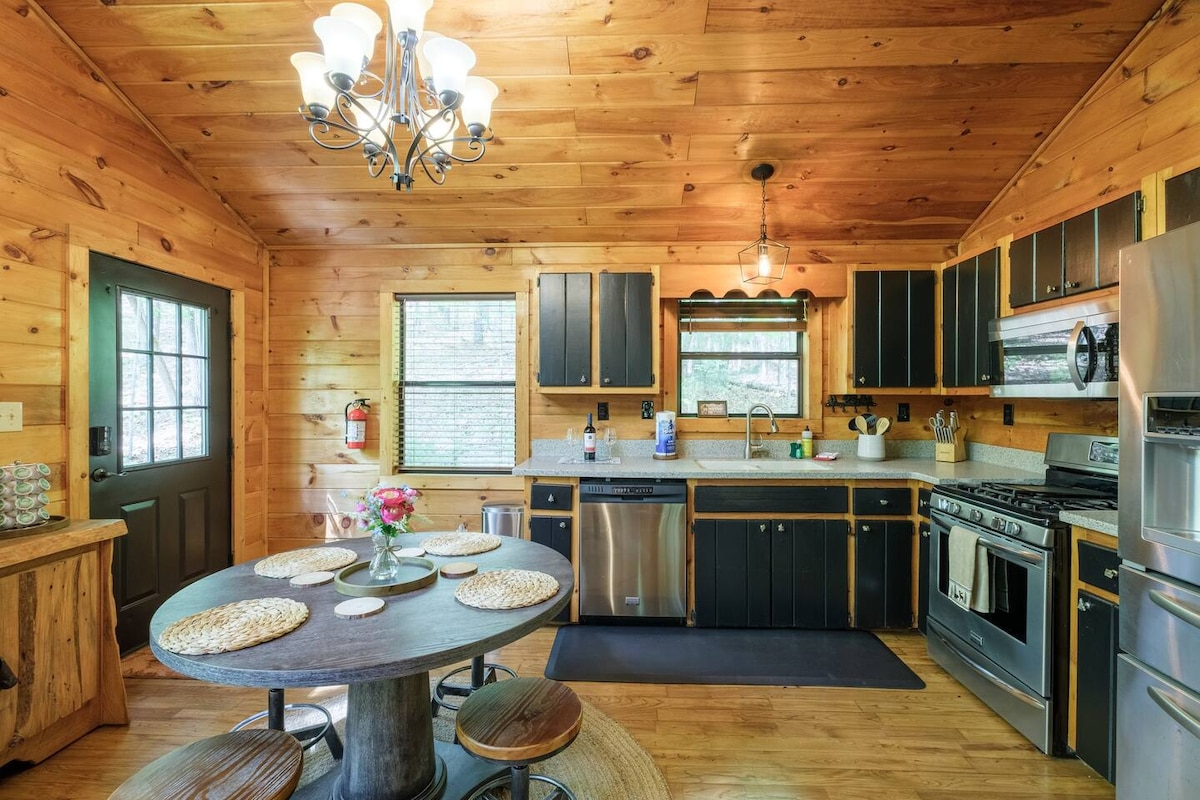 The kitchen showcases wooden cabinetry and a spacious countertop with modern appliances. A round dining table with seating for four is positioned beside a window, allowing natural light to fill the space. Warm wooden accents create an inviting cabin atmosphere.