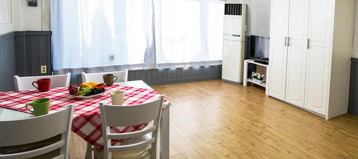 A spacious living area features a dining table set with four chairs and a checkered tablecloth. Large windows allow natural light to fill the room, illuminating the wooden floor. A refrigerator and a small television are visible in the background.