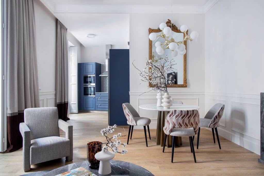 A dining area is illuminated by natural light, featuring a round table surrounded by four upholstered chairs. A contemporary chandelier hangs above, while a large mirror creates a spacious feel. Light wooden flooring complements the neutral color palette, and decorative vases and branches add subtle accents.