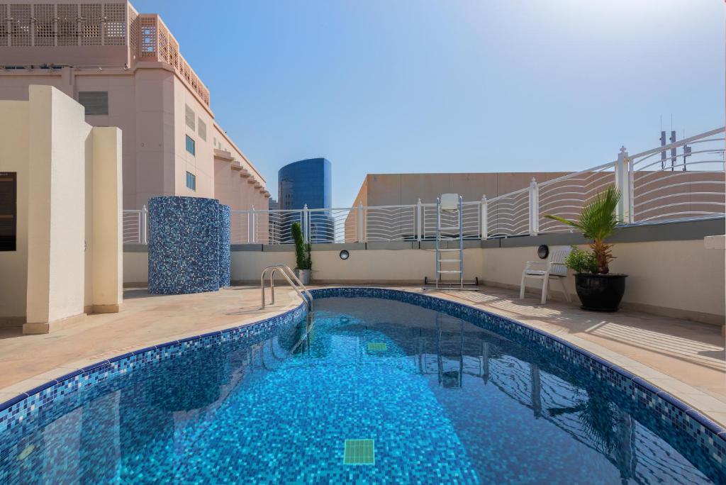 The outdoor pool showcases a tiled surface with varying shades of blue, surrounded by a spacious deck. A ladder provides access to the pool, and a potted plant adds a touch of greenery. A sleek building can be seen in the background against a clear sky.