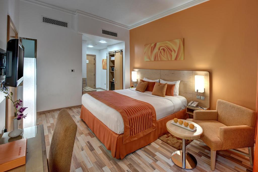 A well-appointed room is shown featuring an inviting spacious bed with coordinating soft furnishings in warm orange tones. A cozy seating area includes an upholstered chair and a small round table. Natural light is allowed in through a partially covered window, enhancing the overall ambiance.