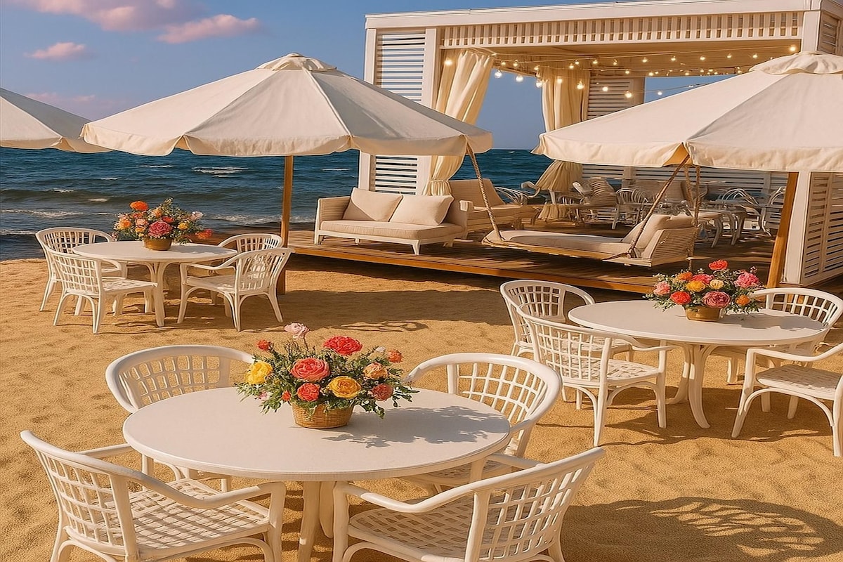 An inviting beachside setup features several white tables surrounded by matching chairs. Each table is adorned with floral arrangements. In the background, a shaded lounge area with comfortable seating is visible, providing a perfect spot to relax while enjoying the seaside view.