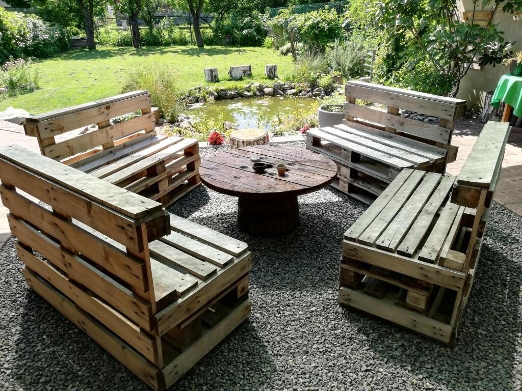 A seating area made from wooden pallets is arranged in a circular formation around a central table, situated on a gravel surface. Lush greenery and a pond can be seen in the background, enhancing the outdoor atmosphere.