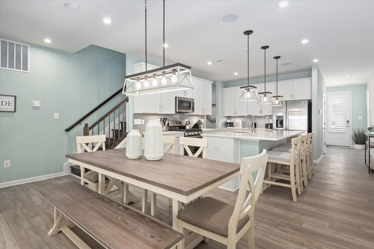 An open-concept kitchen and dining area is highlighted by a large wooden table with seating for ten. A modern kitchen with stainless steel appliances and granite countertops is visible in the background, alongside pendant lighting that enhances the contemporary feel.