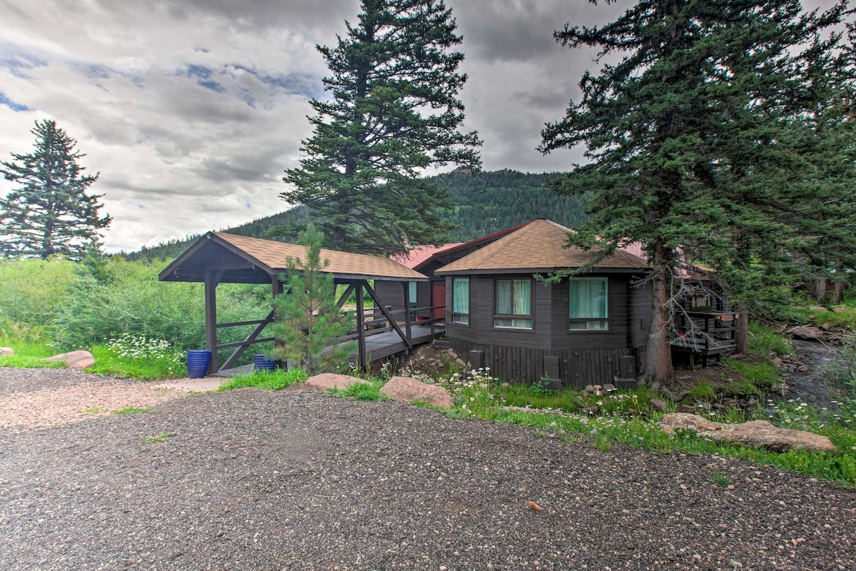 The vacation rental home is nestled in a natural setting, surrounded by tall pine trees and grassy areas. The exterior features a mix of wooden siding and a red roof, with a gravel driveway leading to the entrance. Nearby, the Cucharas River flows gently.