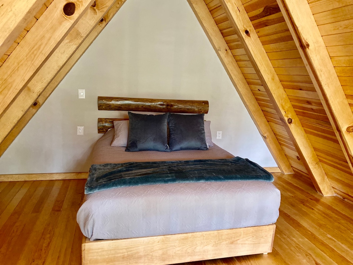 A spacious bed with a wooden frame is positioned under the slanted ceiling, featuring two dark throw pillows and a soft blanket. Natural wood beams and light hardwood flooring create a warm and calming atmosphere.