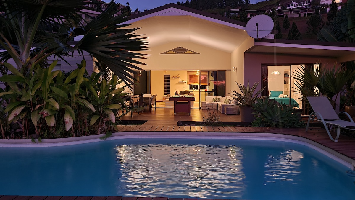 The villa's exterior is illuminated at dusk, featuring a refreshing pool surrounded by tropical plants. In the background, the indoor living space presents an inviting layout, with large glass doors providing a seamless connection to the outdoor area.