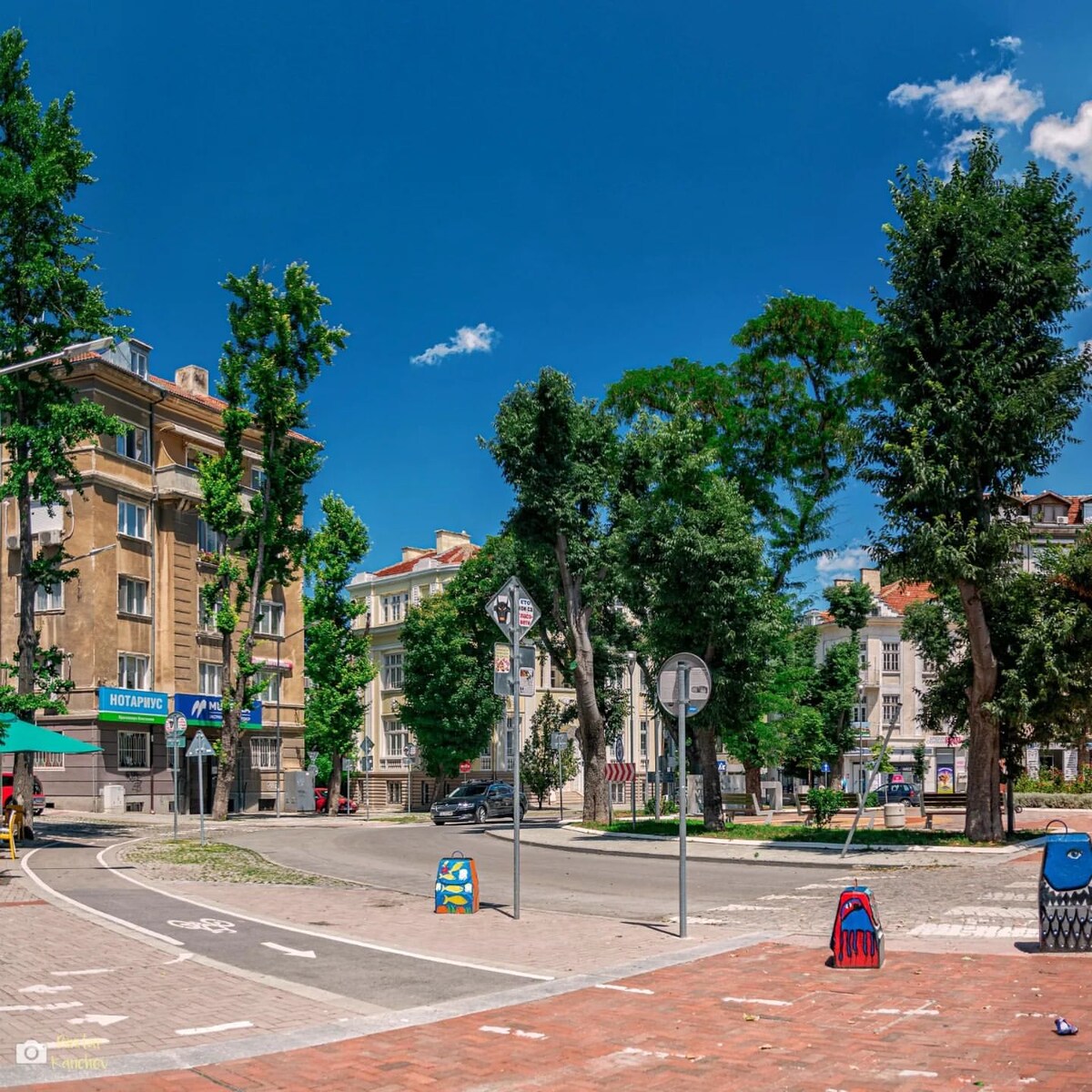 A vibrant street scene captures the lively atmosphere of a central square in Varna. Lush green trees line the road, and colorful character figures add a playful touch. Historic buildings frame the area, with clear blue skies overhead, inviting leisurely exploration of the surroundings.