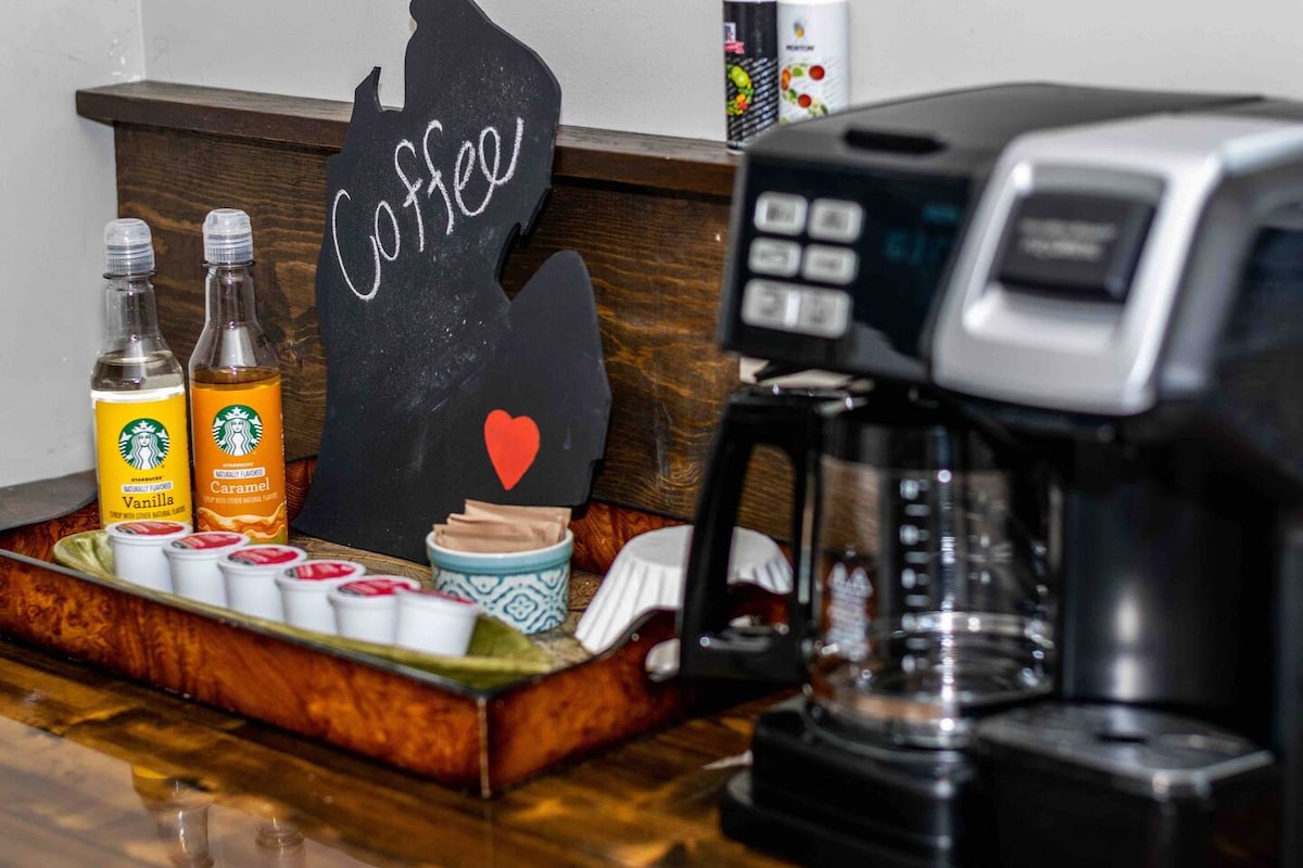 A coffee station is presented on a wooden countertop, featuring a coffee maker and a decorative tray. Bottled flavored syrups are placed next to single-serve coffee pods, with a handwritten sign reading 'Coffee' in the shape of Michigan. Cozy and functional, it invites a warm beverage experience.
