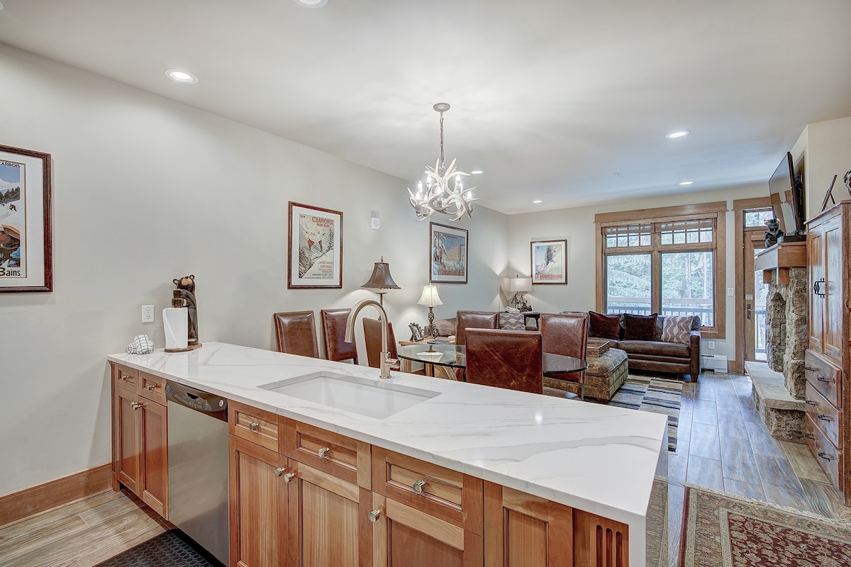 The open living area showcases a modern kitchen island with a white countertop and wooden cabinetry. A dining area is positioned next to the kitchen, while a cozy seating area features a leather sofa and decorative artwork on the walls, with natural light illuminating the space.