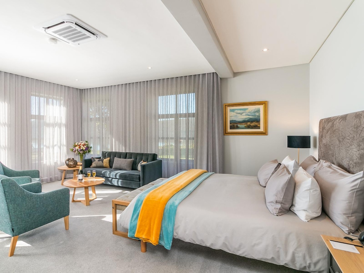 A spacious bedroom features a king size Tempur bed draped in soft linens. A cozy seating area includes a green sofa and two armchairs, supplemented by a coffee table. Floor-to-ceiling curtains allow natural light to filter in, enhancing the serene ambiance.