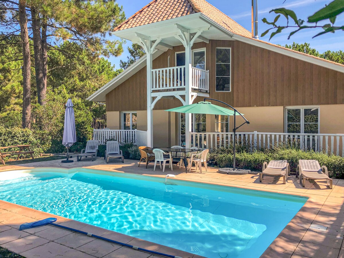 The image features a serene outdoor area with a private swimming pool surrounded by a spacious sun deck. Several lounge chairs are positioned around the pool, and a patio set with an umbrella is visible nearby. The villa's exterior is highlighted by a rustic wooden facade and a balcony overlooking the pool.