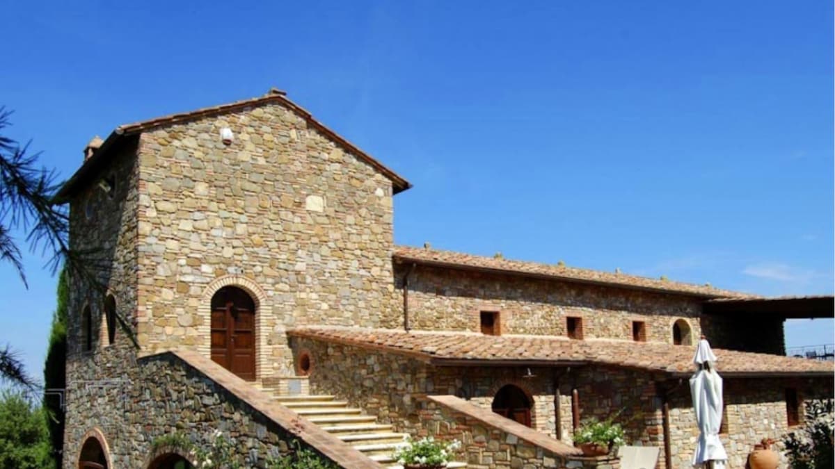 A rustic farmhouse constructed of warm, natural stone is set against a clear blue sky. Stairs lead to an inviting entrance, framed by large arched windows. The structure showcases a traditional tiled roof and has an outdoor seating area with a sun umbrella.