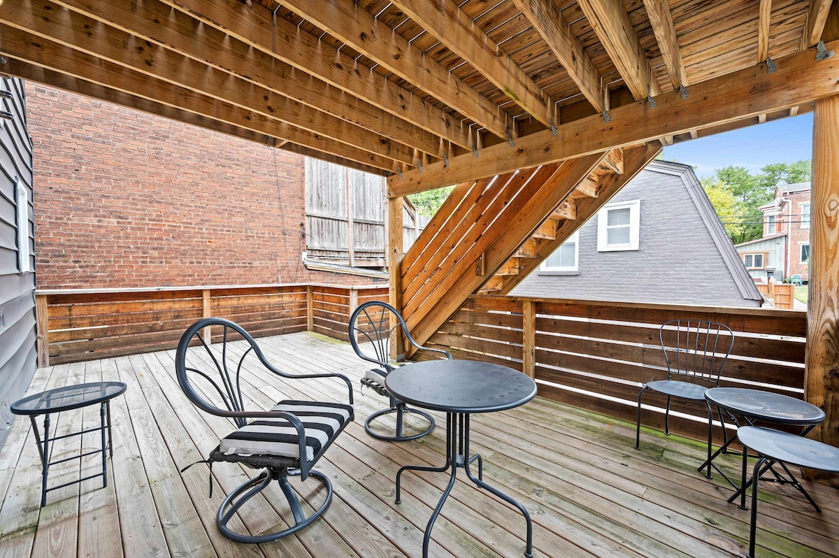 A spacious outdoor deck features wooden flooring and is sheltered beneath upper stairs. Two lounge chairs and a round table invite relaxation, while four additional black wire chairs offer ample seating. The surrounding quiet neighborhood is visible, adding a serene backdrop to the outdoor space.