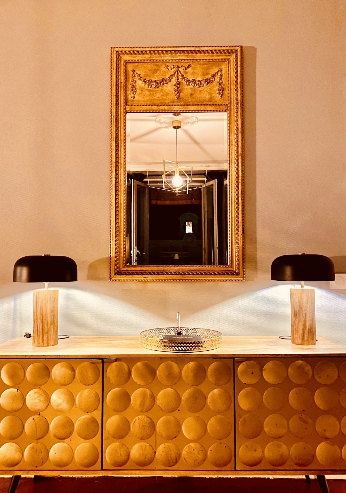 A decorative console table with rounded details is displayed against a neutral wall. Two small black lamps flank a wooden tray on the table. A large, ornate mirror above reflects ambient light and adds depth to the space, creating an elegant focal point.