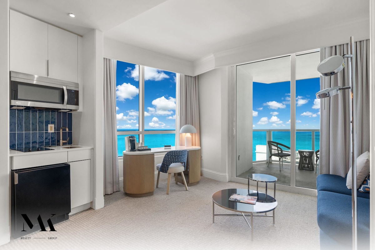 A bright and airy suite is displayed, featuring a kitchenette with stainless steel appliances and a blue-tiled backsplash. A comfortable seating area includes a blue sofa and circular coffee table, with large windows offering stunning ocean views and natural light.