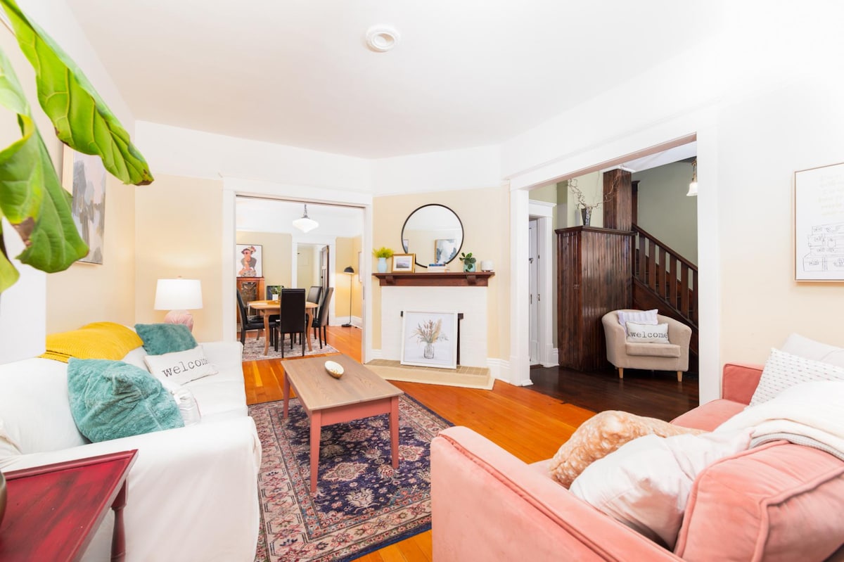 A welcoming living space features a combination of cozy seating with colorful cushions, a polished wooden coffee table, and a decorative area rug. Soft lighting is provided by a floor lamp, and the room connects to a dining area visible through an open doorway.