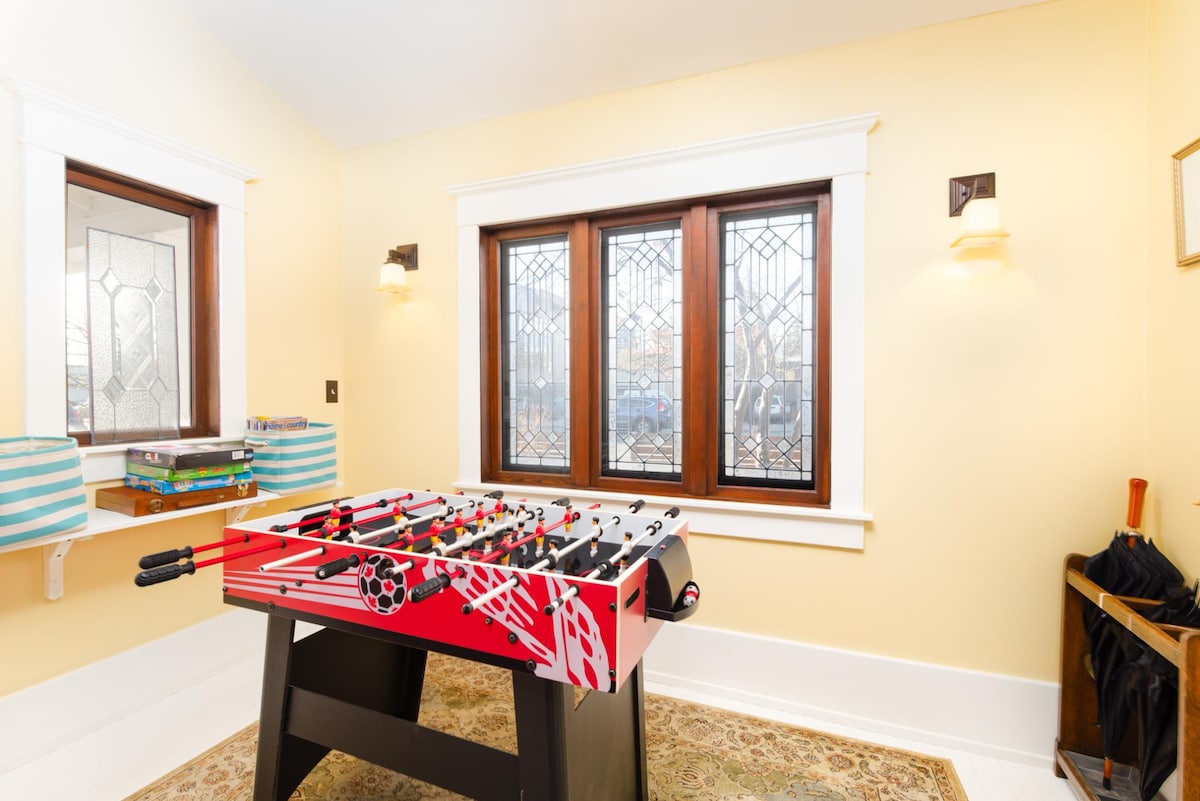 A foosball table with a vibrant red and black design is positioned in a well-lit room. Two large windows featuring decorative stained glass provide natural light, while soft wall sconces add to the illumination. A patterned area rug lies beneath the table, enhancing the cozy ambiance.