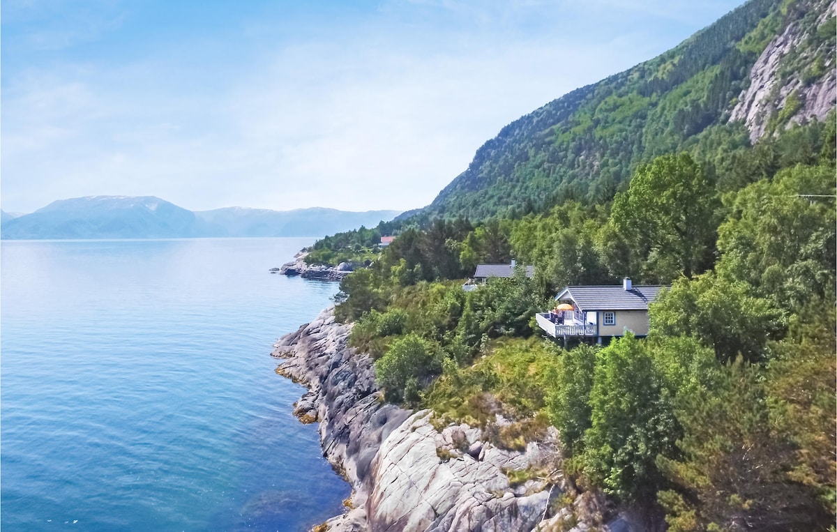 A serene view of the cottage perched along the coastline, showcasing the calm waters of the fjord. Lush greenery surrounds the property, while majestic mountains rise in the distance, complementing the natural landscape.