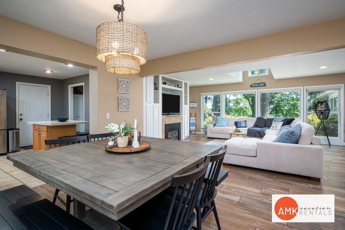 A spacious dining area features a large wooden table surrounded by black chairs. Natural light fills the room, highlighting an open floor plan that connects to a cozy living space with a large sectional sofa and expansive windows overlooking the outdoors.