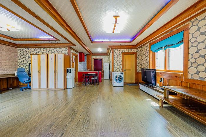 An airy, open space features wooden accents and a light-colored floor. Various furniture pieces include a dining table, a chair, a refrigerator, and a television. Natural light enters through the windows, enhancing the warm atmosphere of the room.