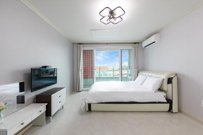 A well-lit bedroom is furnished with a spacious bed and a television mounted on the wall. Large glass doors provide access to an outdoor area, allowing natural light to fill the room. Light-colored walls and simple decor create a clean and modern atmosphere.