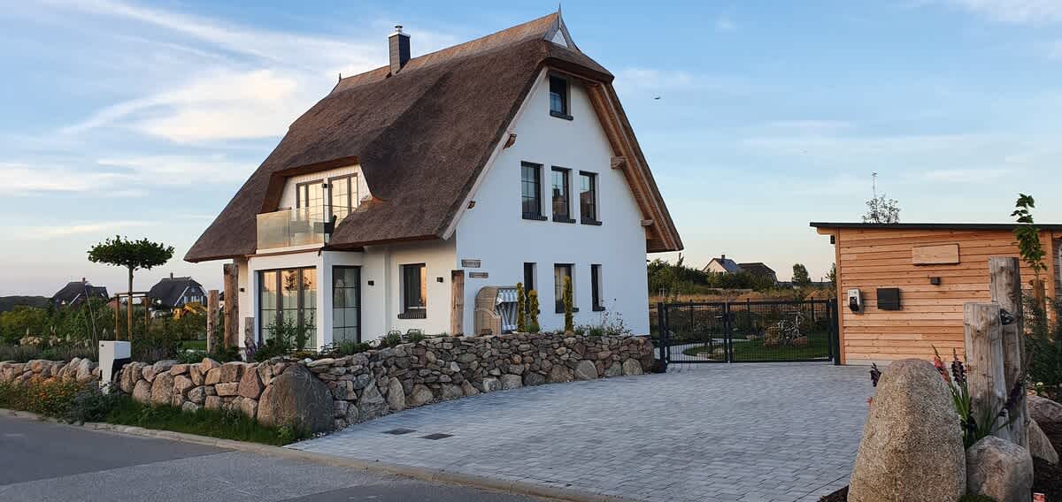 A modern thatched-roof holiday home is positioned amidst a rural landscape, featuring a stone wall and a gated entrance. The structure showcases a blend of white walls and wooden details, providing a contemporary look with large windows offering views of the surroundings.