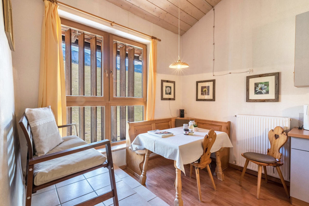 The cozy dining area features a wooden table surrounded by elegantly carved chairs. A comfortable armchair is positioned nearby. Natural light flows through large windows, adorned with light curtains, offering views of the landscape beyond. Framed artwork adorns the walls, adding a touch of charm.