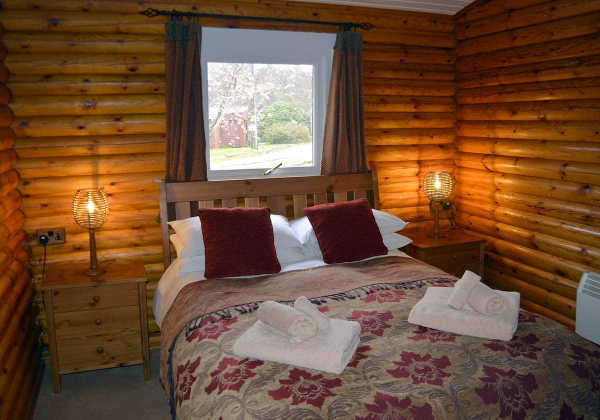 A double bedroom features a 4'6" bed adorned with crisp white linens and decorative pillows. Two bedside tables flank the bed, each with a lamp providing soft lighting. A window lets in natural light, and coordinated curtains frame the view of the surrounding greenery.
