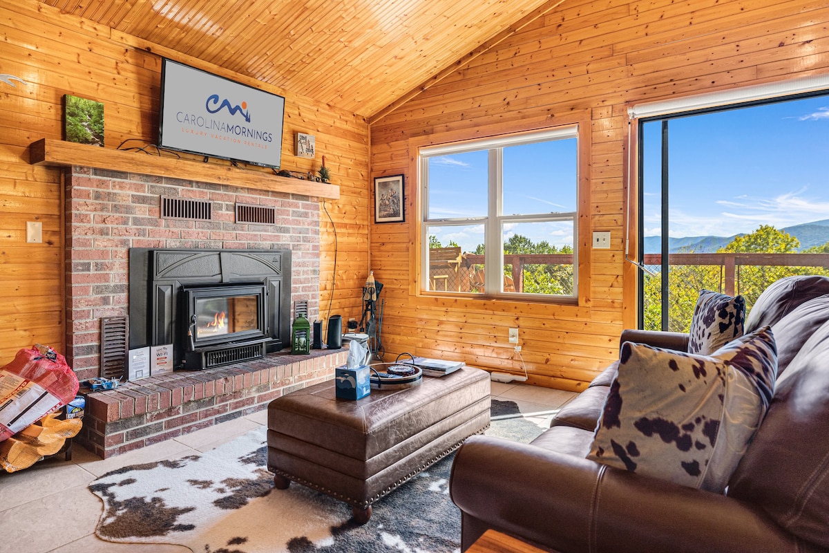 A warm and inviting living area features wood-planked walls and ceiling, showcasing a comfortable sofa with patterned pillows. A wood stove is centered beneath a television, while a large window reveals expansive mountain views. A coffee table sits in front of the seating arrangement.