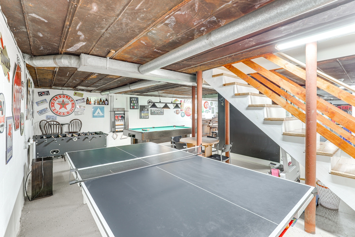 A spacious game room is featured, showcasing a black ping pong table with ample space for play. Vintage wall art and signage adorn the white walls. In the background, stairs lead to the upper level, with wooden steps highlighted by industrial fixtures.
