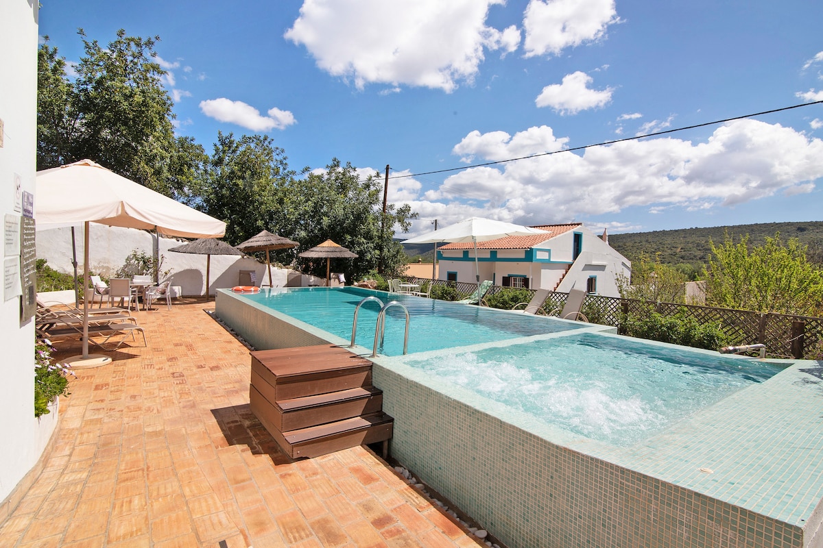The outdoor area features a large swimming pool with steps leading into the water. The surrounding patio is lined with sun loungers and umbrellas, with greenery visible in the background, creating a serene environment for relaxation.