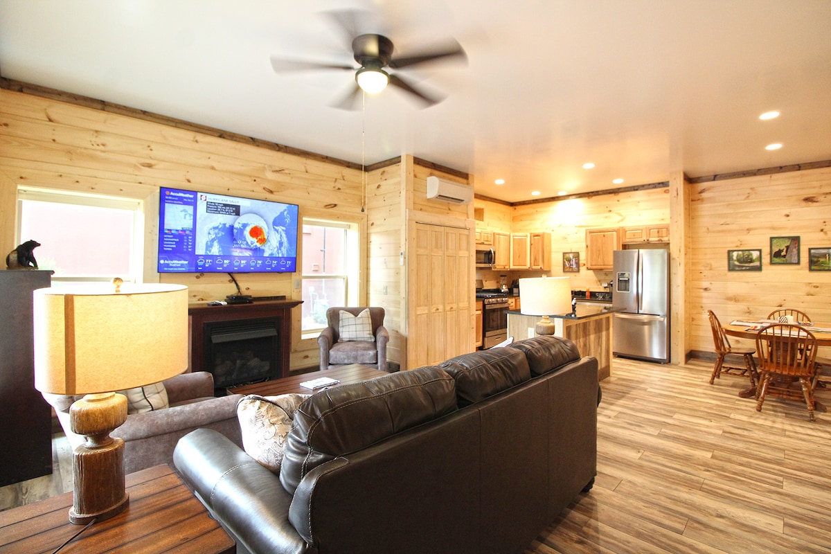 An open living area showcases hardwood flooring and natural wood paneling. A comfortable sofa is positioned near a gas fireplace, while a wall-mounted flatscreen TV provides entertainment. The kitchen features stainless appliances, with a dining area that includes a wooden table and chairs.