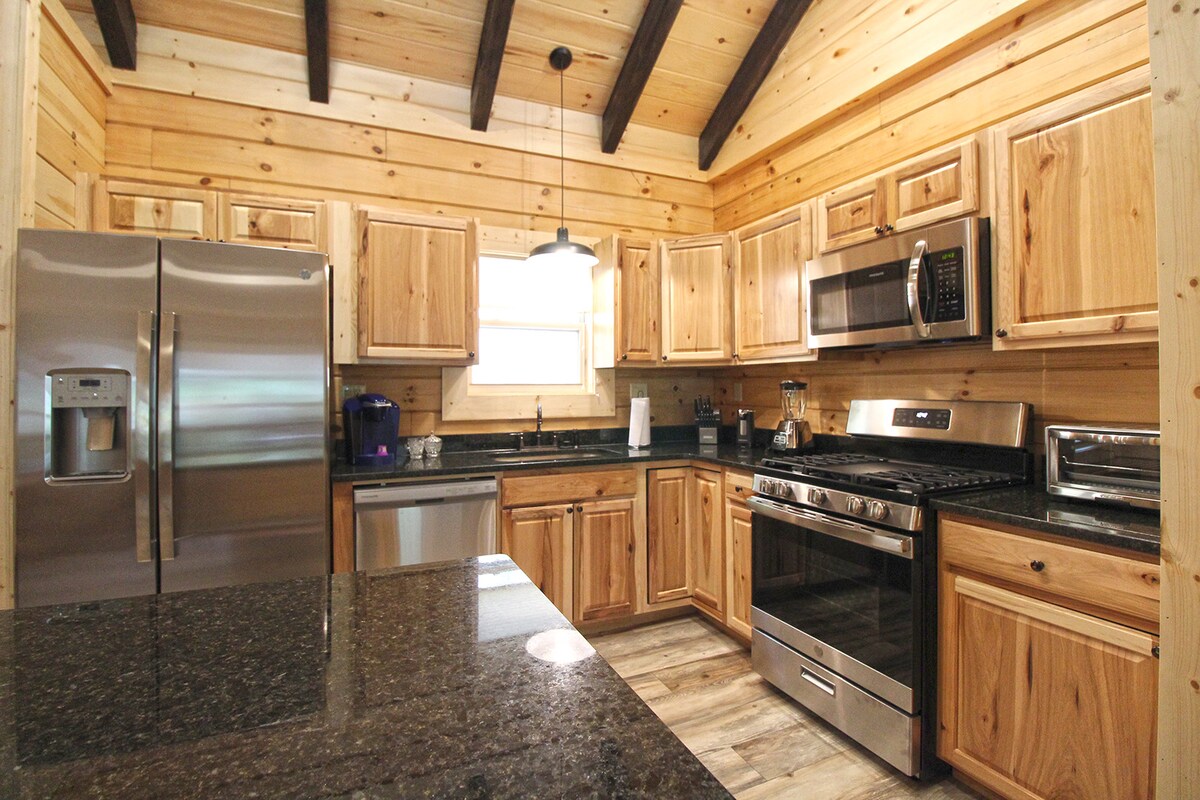 A kitchen is showcased with wooden cabinetry and modern stainless appliances, including a refrigerator and gas range. The granite countertops complement the rustic wood paneling and exposed beams above. Natural light enters through a window, illuminating the workspace.