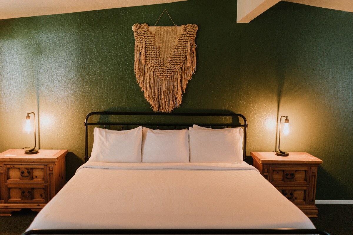 A spacious bed is positioned centrally in the room, flanked by wooden nightstands. Soft lighting from bedside lamps illuminates the area. A textured wall hanging adds visual interest, while rich green walls create an inviting backdrop.