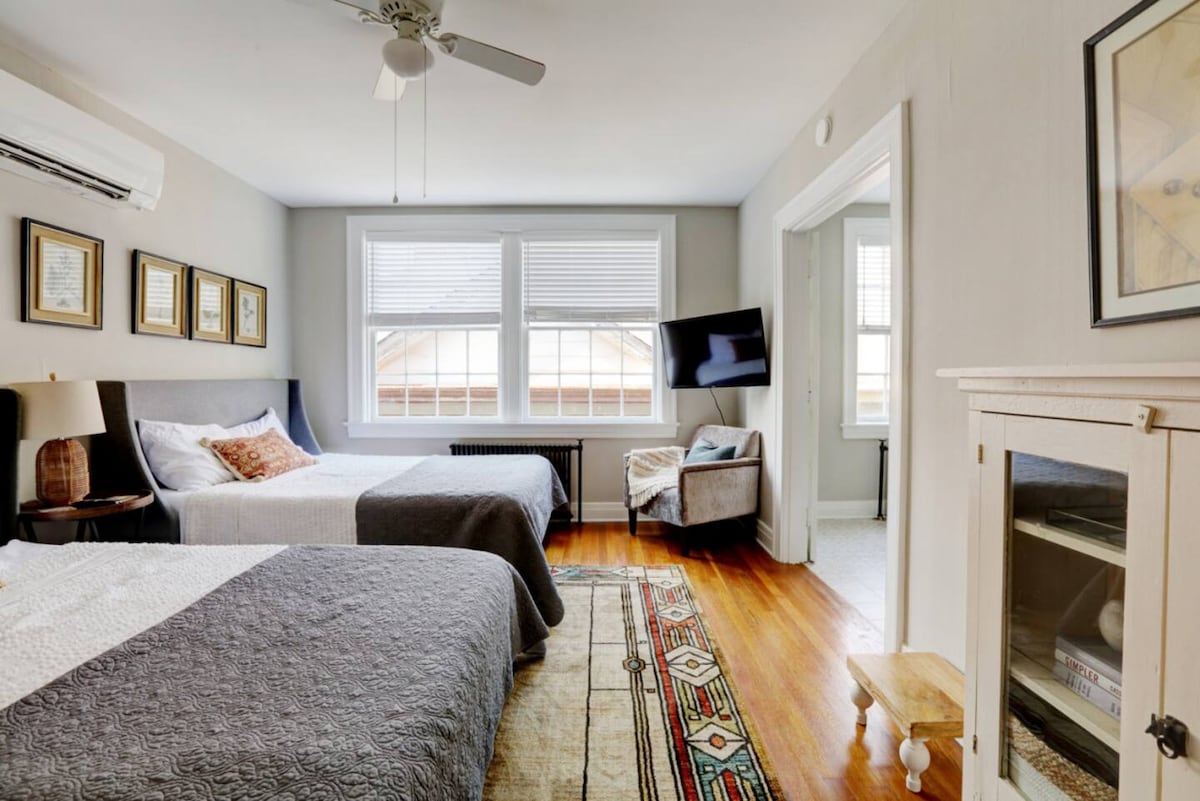 A spacious room is equipped with two double beds, soft grey bedding, and decorative wall art. A comfortable armchair is positioned near a wall-mounted television. Large windows allow natural light to fill the space, while a ceiling fan adds to the room's comfort.