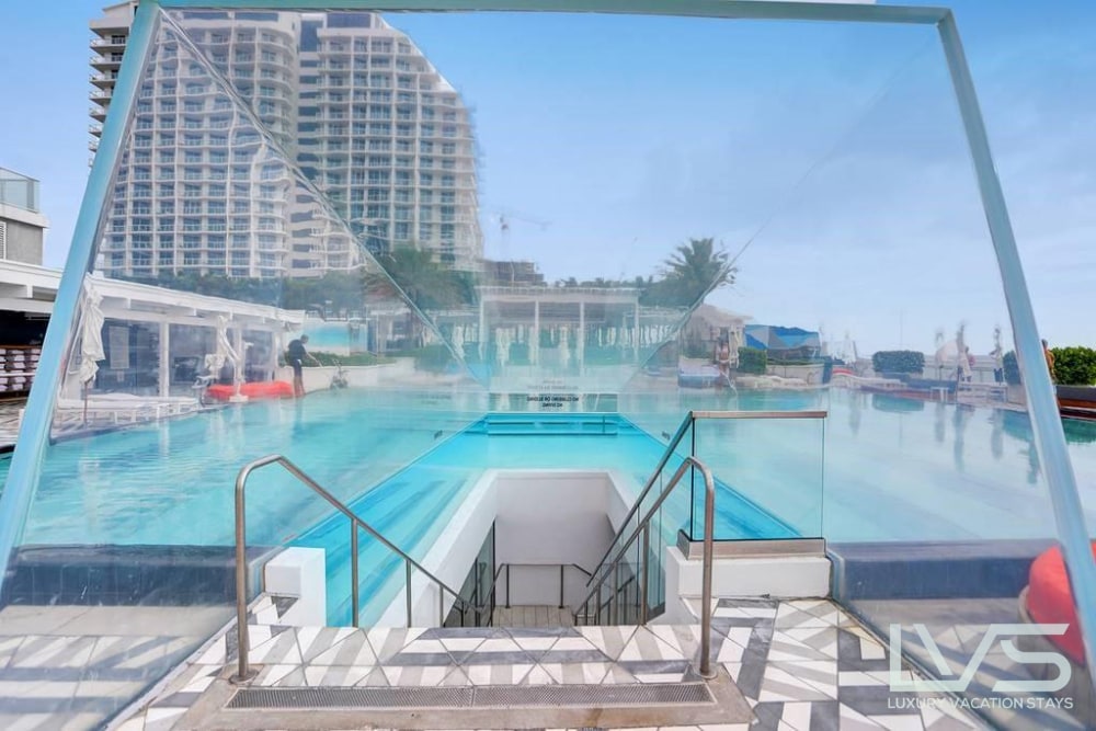 A modern pool area features a clear glass structure leading down to the water. Lounge chairs are arranged around the perimeter, while a spacious swimming pool reflects the surrounding palm trees and architecture, creating a serene ambiance.